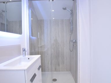A bathroom with a walk in shower with wooden panel walls and a white sink cabinet at Sea Green Cottage Walberswick