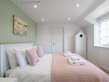 A bedroom with a bed with pink pillows and blanket a painting on a green wall and a white wardrobe by a window at Sea Green Cottage Walberswick
