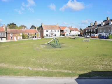 A grassy area with a playground slide and swings surrounded by houses and benches at Sea Green Cottage Walberswick