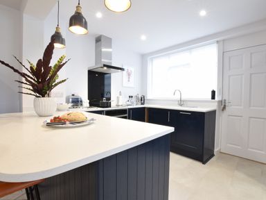 A kitchen with dark blue cabinets white countertops a white door and a window with blinds at Sea Green Cottage Walberswick