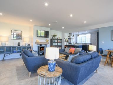 A living room with blue sofas a wooden side table with a lamp and a kitchen with pendant lights in the background at Sea Green Cottage in Walberswick