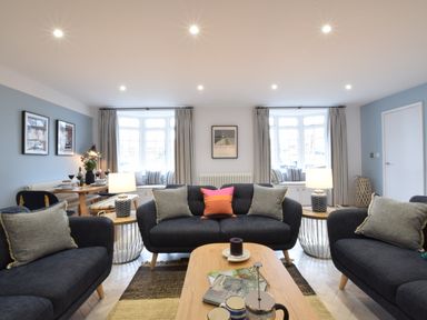 A living room with dark sofas, a wooden coffee table, side tables with lamps, and dining area with table and chairs at Sea Green Cottage Walberswick