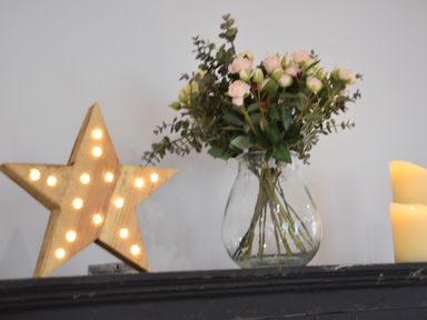A wooden lighted star decoration next to a glass vase with flowers and a lit candle on a black surface at Sea Green Cottage Walberswick