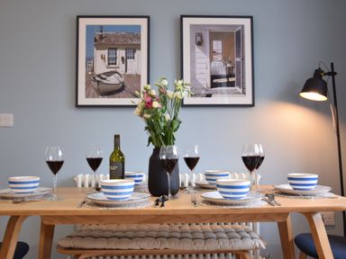 A dining table set with blue striped bowls wine glasses and a vase of flowers with framed pictures on the wall and a floor lamp at Sea Green Cottage Walberswick