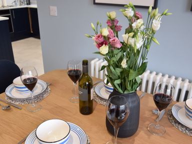 A dining table set with four plates bowls utensils glasses of wine a bottle of wine and a vase of flowers in a kitchen at Sea Green Cottage Walberswick