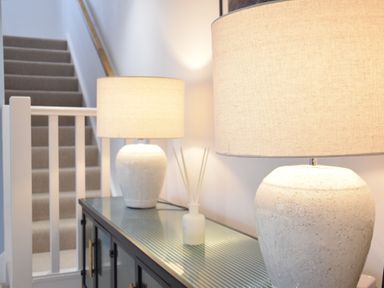 A hallway with a black cabinet topped with two white lamps and a diffuser next to a carpeted staircase at Sea Green Cottage Walberswick