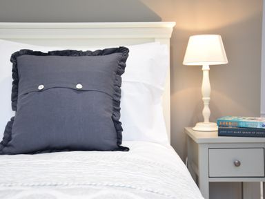 A bed with a large dark pillow white bedding and a bedside table with a lamp and books at Sea Green Cottage Walberswick