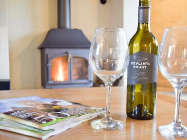 A table with two empty wine glasses a bottle of Devlin's Mount Shiraz wine two magazines and a wood burning stove in the background at Church Farmhouse Cookley Near Halesworth