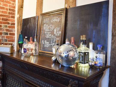 A wooden bar countertop with bottles, disco balls, a framed cocktail menu, a lava lamp, and a radio at Church Farmhouse Cookley Near Halesworth