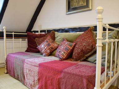 A room with a white metal framed daybed covered in a red patterned throw and assorted decorative cushions at Church Farmhouse Cookley Near Halesworth