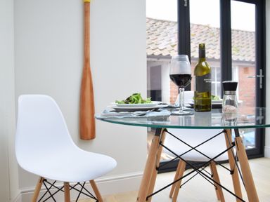 A dining area with a glass table and two white chairs with wooden legs with a bottle of wine wine glasses plates of salad and a salt grinder at Arlo Cottage Orford