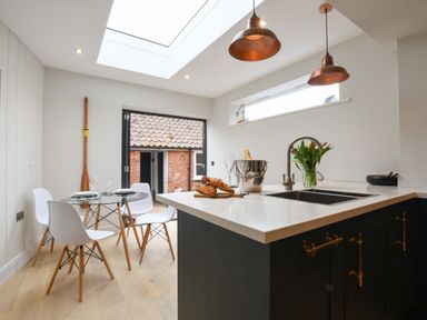 A kitchen with a glass dining table and white chairs next to a sliding door and an island countertop with a sink and croissants at Arlo Cottage Orford