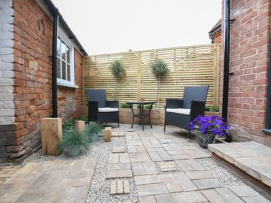 A small outdoor patio with two black chairs and a round table between brick walls and wooden fence at Arlo Cottage Orford