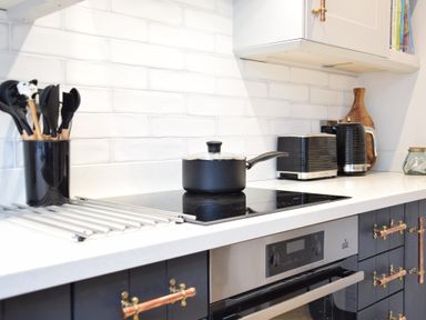 A kitchen countertop with cooking utensils in a container a saucepan on an electric stovetop a toaster and a kettle with white tiled backsplash at Arlo Cottage Orford