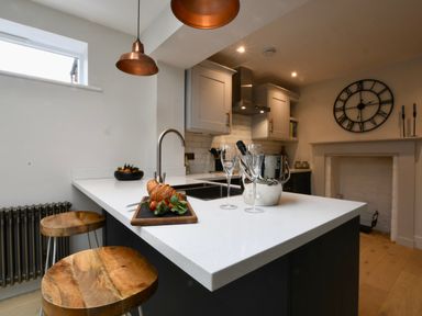 A kitchen with a white countertop island with croissant and strawberries on a cutting board two wooden stools and a wall clock at Arlo Cottage Orford