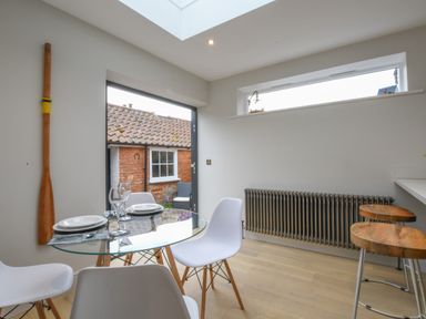 A dining area with a glass table and white chairs next to a door showing an outdoor view with a brick building at Arlo Cottage Orford