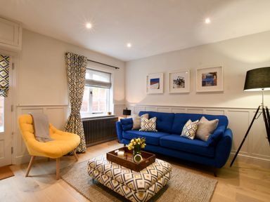 A living room with a blue sofa yellow chair patterned curtains and ottoman and a floor lamp at Arlo Cottage Orford
