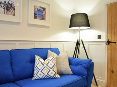 A blue sofa with two cushions next to a black tripod floor lamp and a wooden door at Arlo Cottage Orford
