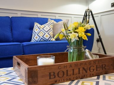 A blue sofa with patterned cushions and a wooden tray holding a jar of yellow flowers and a white candle at Arlo Cottage Orford