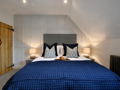 A bedroom with a bed with blue and patterned pillows a blue blanket two bedside tables with lamps and a wooden door at Arlo Cottage Orford