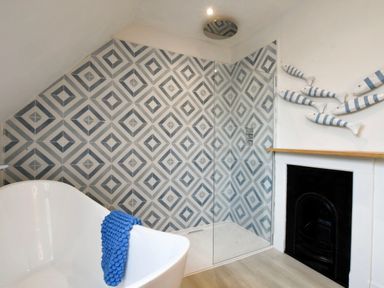 A bathroom with a freestanding bathtub a blue towel a glass shower enclosure with patterned tiles and a fireplace with fish wall decor at Arlo Cottage Orford