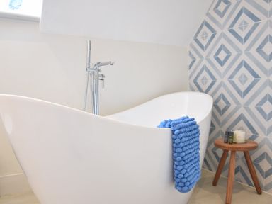 A white freestanding bathtub with a blue towel and a wooden stool with toiletries against a wall with blue and white patterned tiles at Arlo Cottage Orford