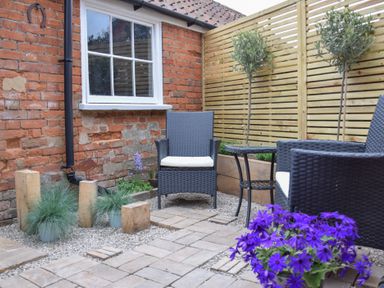An outdoor patio with two chairs a small table brick wall wooden fence and purple flowers at Arlo Cottage Orford