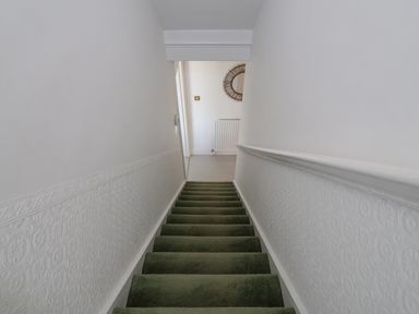 A staircase with green carpet and white textured walls leading to a landing with a radiator and round mirror at Time Out Southwold in Southwold