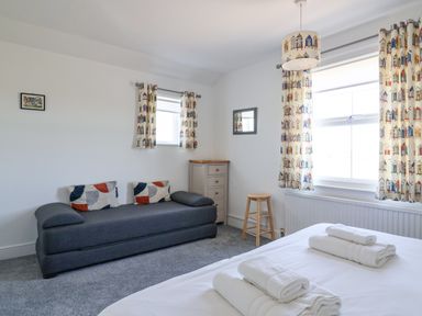 A bedroom with a bed with folded towels a gray sofa with cushions a wooden stool a chest of drawers curtains and a pendant light at Time Out Southwold in Southwold