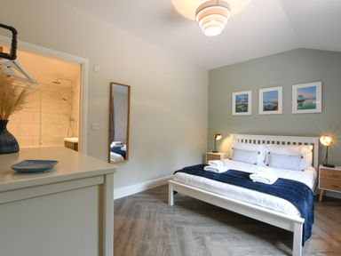 A bedroom with a white bed and blue blanket with bedside tables and lamps and a bathroom visible at The Piglets in Newbourne Near Woodbridge