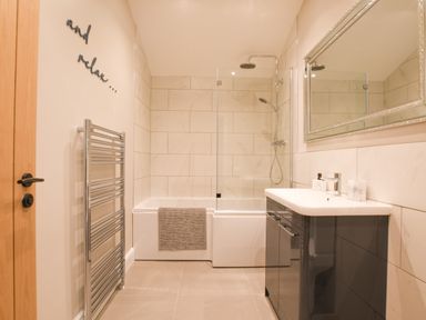 A bathroom with a bathtub shower combo a towel radiator a sink with storage cabinets and a mirror at The Piglets in Newbourne Near Woodbridge
