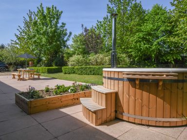 A wooden hot tub with steps next to a raised garden bed and a patio table with benches and umbrella in a garden area at The Piglets in Newbourne Near Woodbridge