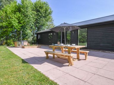 An outdoor patio area with a wooden picnic table and benches under an umbrella near a black building at The Piglets in Newbourne Near Woodbridge