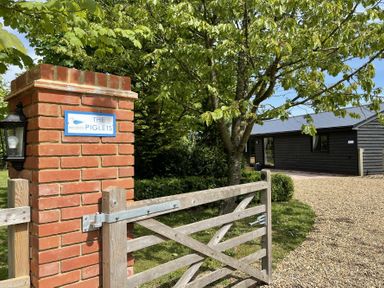A wooden gate and brick pillar with a sign reading The Piglets near a tree and black building with gravel driveway at The Piglets in Newbourne Near Woodbridge