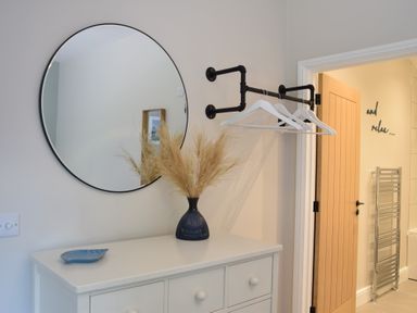A white dresser with a black vase of dried plants underneath a round mirror and three white clothes hangers on a black wall rack at The Piglets in Newbourne Near Woodbridge