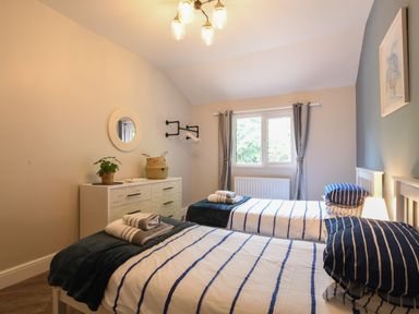 A bedroom with two single beds a dresser a round wall mirror a window with curtains and a ceiling light at The Piglets in Newbourne Near Woodbridge