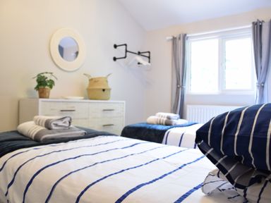 A bedroom with two single beds with striped bedding towels on beds dresser with plant and basket wall mirror and window with curtains at The Piglets in Newbourne Near Woodbridge