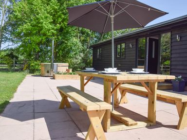 An outdoor patio with a wooden picnic table and benches under a large umbrella near a dark wooden building at The Piglets in Newbourne Near Woodbridge