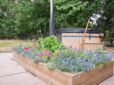 A wooden raised flower bed with various blooming flowers and greenery next to a wood-fired hot tub in a garden setting at The Piglets in Newbourne Near Woodbridge