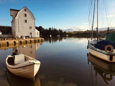 A small motorboat named Isla and a sailboat docked near a white wooden building by calm water at The Piglets in Newbourne Near Woodbridge