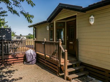 The exterior entrance of a house with wooden steps and railing and a covered grill on a paved area at The Hide Out in Aldeburgh