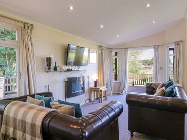 A living room with two leather sofas, a wall-mounted TV, a fireplace, and large windows with curtains at The Hide Out, Aldeburgh