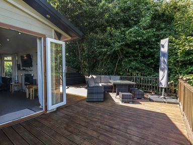 An outdoor wooden deck with a wicker sofa set and table next to a building with an open glass door at The Hide Out in Aldeburgh