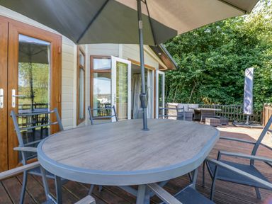 An outdoor deck with a table and chairs under an umbrella and lounge seating surrounded by trees at The Hide Out Aldeburgh