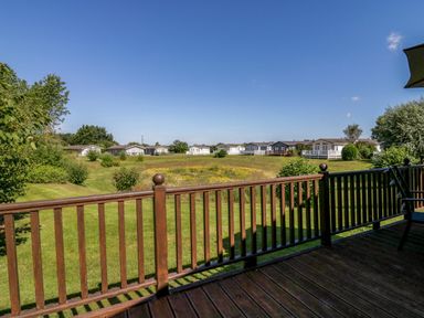 A wooden deck with railing overlooking a grassy area with bushes and white houses at The Hide Out in Aldeburgh