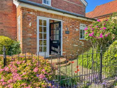 A brick cottage entrance with an open black door and white-framed windows surrounded by flowers and a black metal fence at Chapel Cottage Newbourne Near Woodbridge