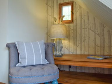 A corner with a gray armchair with a cushion a wooden bench with a rabbit shaped lamp and magazines on it at Chapel Cottage Newbourne Near Woodbridge