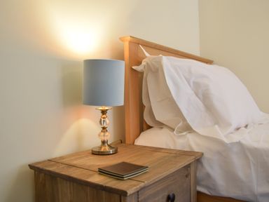 A bedside table with a lamp and notebooks next to a bed with white pillows at Chapel Cottage Newbourne Near Woodbridge