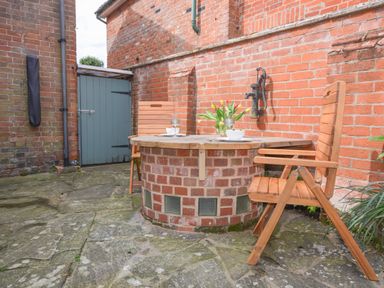 A small outdoor patio area with a round brick table with a wooden top two wooden chairs a vase with flowers on the table and a green wooden door at Chapel Cottage Newbourne Near Woodbridge