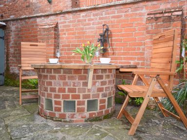 An outdoor seating area with a round brick table wooden chairs and a flower vase against a brick wall at Chapel Cottage Newbourne Near Woodbridge